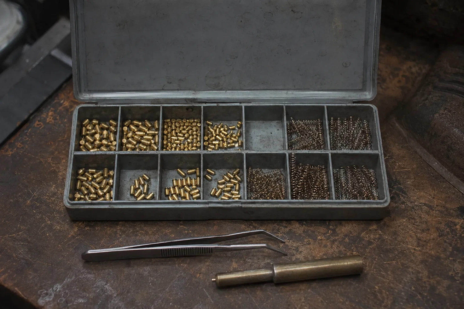Open metal case with brass pins and coiled springs sorted in small sections, with tweezers and a brass tool in front.