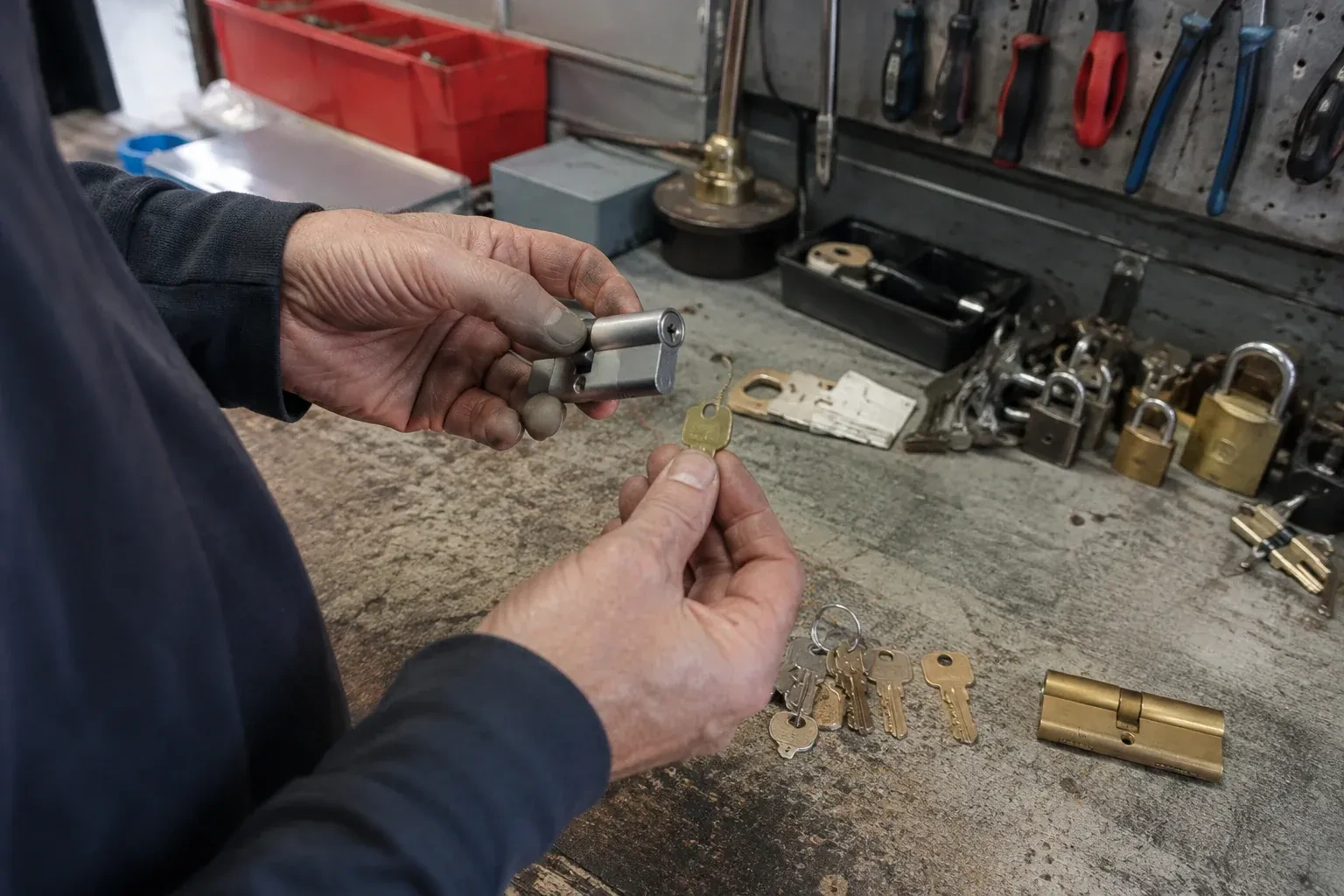 Hands holding a silver-colored lock cylinder and a brass-colored key above a workbench with padlocks, keys and tools. — Master Key System Design, Woodstock
